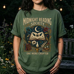 Può includere: Una t-shirt verde oliva con una stampa grafica che riporta il testo "MIDNIGHT READING SOCIETY" e "ONE MORE CHAPTER". Il design include un libro aperto con un occhio, una luna crescente, fiori, una lampada a olio accesa e una penna d'oca.