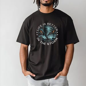 Könnte beinhalten: Schwarzes T-Shirt mit einer Grafik eines Tornados und dem Text "LIFE IS BETTER IN THE STORM". Das Design zeigt einen wirbelnden blauen Tornado mit Blitzen und eine stürmische Landschaft.