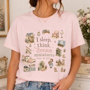 May include: A light pink t-shirt featuring a whimsical illustration of miniature dollhouse furniture and the text "I sleep, think and dream miniatures". The design includes a bed, armchair, and a dollhouse.