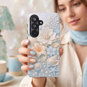 May include: A light blue phone case featuring a 3D floral and seashell design. The case has white, peach, and light blue flowers, seashells, and pearls. A black camera module is visible at the top.