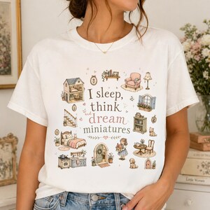 May include: Cream-colored t-shirt featuring the text "I sleep, think and dream miniatures" with illustrations of miniature dollhouse furniture and accessories. The design includes a dollhouse, bed, and kitchen.