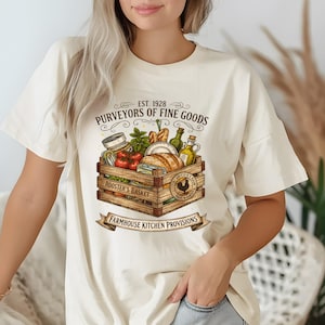 May include: Cream-colored t-shirt featuring a graphic of a wooden crate filled with food items like tomatoes, bread, and olive oil. The text on the shirt reads "PURVEYORS OF FINE GOODS" and "ROOSTER'S BASKET", with "FARMHOUSE KITCHEN PROVISIONS" on a banner.