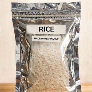 May include: A silver foil bag filled with white rice, labeled "RICE" and "MADE IN USA 04/2020." The bag is sealed and ready for long-term food storage. Text at the top reads "Long-Term Food Storage."