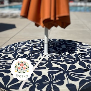 May include: A white and navy blue round tablecloth with a floral pattern. The tablecloth has a zipper around the edge and a hole in the center for an umbrella pole. The tablecloth is branded with the word "fresko" in a circle with a floral design.
