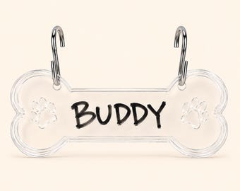 May include: A clear, bone-shaped acrylic pet tag featuring the name BUDDY printed in black lettering. The tag includes two etched paw prints, one on each side of the name, and is attached to two silver-colored metal hooks for hanging purposes.