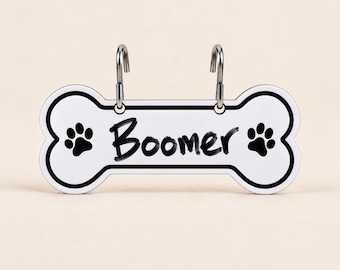 May include: A white bone-shaped pet tag with a black border and two black paw prints on either side. The name Boomer is printed in black script in the center. Two silver metal hooks are attached to the top of the tag for hanging purposes.