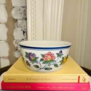 May include: A white ceramic bowl with a blue and white floral design. The bowl is sitting on top of three books. The top book is yellow and says "A Bouquet of Roses". The middle book is pink and says "I know I am, but what are you?" The bottom book is blue and says "From Paycheck to Purpose".