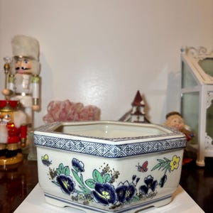 May include: A white, hexagonal ceramic planter with a blue patterned rim and floral design. The planter features blue and yellow flowers with green leaves and small insects. The planter sits on a book with the word "SUPERSIGHT" in red.