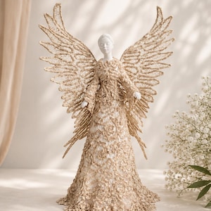 Puede incluir: Una figura decorativa de ángel con una estructura base blanca adornada con intrincados detalles de encaje de color beige en la túnica y grandes alas extendidas. La figura tiene un rostro y manos blancas lisas, con encaje de textura detallada.