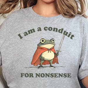 May include: A light gray t-shirt with the text "I am a conduit FOR NONSENSE." The graphic features a cartoon frog wearing a red cape and holding a sword. The frog has large eyes and a cream-colored belly.