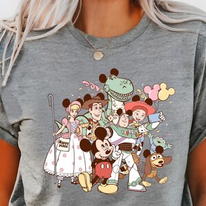 May include: Gray t-shirt featuring a colorful graphic of Disney characters, including Mickey Mouse, Buzz Lightyear, and Woody. The design includes balloons and confetti, creating a festive and playful vibe. The shirt is made of a soft material.