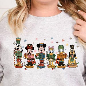 May include: A light gray sweatshirt featuring a line of Disney characters designed as nutcrackers. The characters include Mickey Mouse, Minnie Mouse, Donald Duck, Daisy Duck, Goofy, and Pluto, all in festive outfits. Decorative snowflakes and stars are also present.