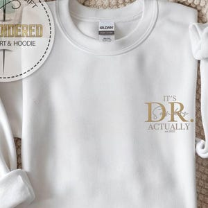 May include: White sweatshirt with the text "IT'S DR. Actually" in gold lettering. The sweatshirt has a crew neck and long sleeves. The design includes the year "est. 2025". The sweatshirt is a personalized gift.