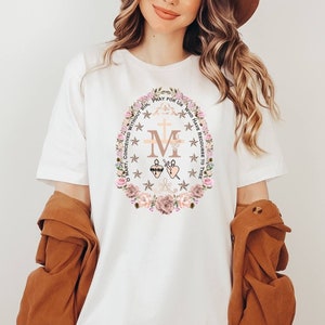 May include: White t-shirt featuring a floral design with the letter "M", a cross, and the words "PRAY FOR US". The design is surrounded by a floral wreath with pink and beige flowers. The person is wearing a brown hat and a brown jacket.