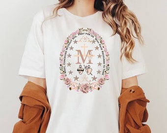 May include: White t-shirt featuring a floral design with the letter "M", a cross, and the words "PRAY FOR US". The design is surrounded by a floral wreath with pink and beige flowers. The person is wearing a brown hat and a brown jacket.