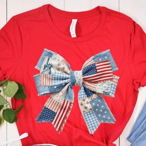 May include: A bright red short-sleeved t-shirt featuring a large, central graphic of a decorative bow. The bow is composed of various fabric patches in red, white, and blue, including patterns of stars, stripes, gingham, and the Statue of Liberty.