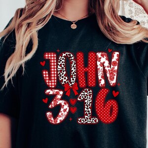 May include: Black t-shirt with the text "JOHN 3:16" in red and white patterns. The word "JOHN" is in a mix of gingham, leopard print, and floral designs. The numbers "3:16" are in floral, leopard print, and polka dot patterns.