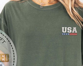May include: Olive green t-shirt with rolled-up sleeves. The shirt features the word "USA" embroidered in gray with a red and blue stripe and stars design. The shirt is made of a soft, comfortable material.