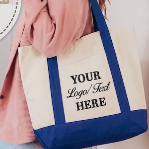 May include: A large off-white canvas tote bag with a solid blue bottom panel and matching blue shoulder straps. The front of the bag features the black printed text YOUR Logo/Text HERE in a combination of serif and script fonts.
