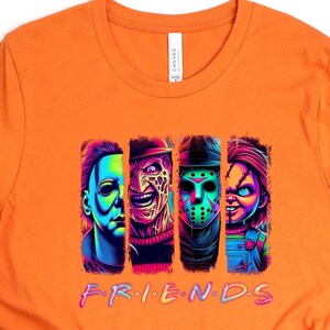 May include: Orange t-shirt featuring a graphic of four horror movie characters in vertical panels. The word "FRIENDS" is written in a colorful, stylized font below the characters. The shirt is made of a soft material.