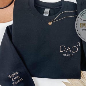 May include: Black sweatshirt with embroidered text "DAD est. 2010" in a light brown thread. The sleeve features the names "Sophia, Emily, James" in the same thread. A gold necklace with a pendant is draped on the sweatshirt. The sweatshirt is folded.