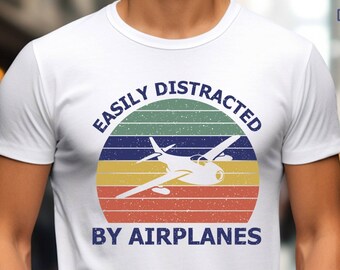May include: White t-shirt with a vintage-style graphic that says "Easily Distracted by Airplanes". The graphic features a plane flying in front of a sunset with a rainbow color scheme.
