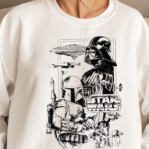 May include: White crewneck sweatshirt featuring a black and white Star Wars graphic. The design includes Darth Vader, Boba Fett, and the Star Wars logo, with various spacecraft and characters.