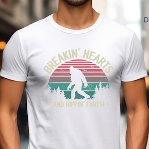 May include: White t-shirt with a Bigfoot graphic inside a retro sunset design. The text reads "BREAKIN' HEARTS" above the image and "AND RIPPIN' FARTS!" below. The shirt is a crew neck style.