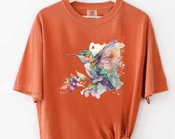 May include: A burnt orange t-shirt with a watercolor hummingbird design. The hummingbird is flying with its wings spread and is surrounded by flowers. The design is in shades of blue, green, purple, and pink.