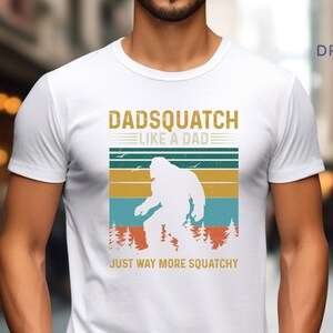 May include: White t-shirt with a retro-style graphic of a Bigfoot with the text "Dadsquatch Like a Dad Just Way More Squatchy".