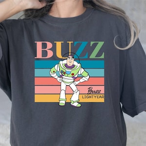 May include: A dark gray t-shirt with a retro-style graphic of Buzz Lightyear from the Toy Story franchise. The graphic features Buzz standing with his arms crossed, and the text "Buzz Lightyear" is printed below him. The text "BUZZ" is printed in large, colorful letters above the graphic.