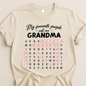 May include: Beige t-shirt with the text "My favorite people call me GRANDMA" in black script. A pink-outlined word search includes names like Olivia, Robert, and David. The shirt is made of a soft material.