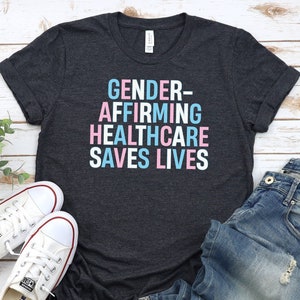 May include: Dark heather gray t-shirt with the text "GENDER-AFFIRMING HEALTHCARE SAVES LIVES" in white letters with pink and blue accents.