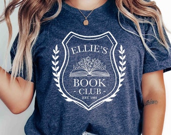 May include: A navy blue t-shirt with a white graphic of a book club logo. The logo features a shield with the text "Ellie's Book Club Est. 1991" and a stylized image of an open book with flowers.