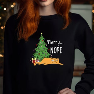 May include: A black sweatshirt with a green Christmas tree and a grumpy orange cat lying under it. The text "Merry...NOPE" is printed on the sweatshirt.
