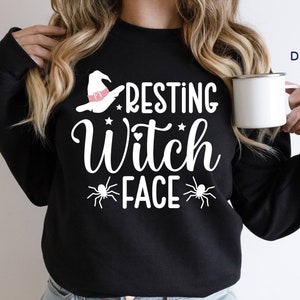 May include: Black sweatshirt with the text "RESTING WITCH FACE" in white, featuring a witch hat and spider graphics. A white mug is held in the hand. The sweatshirt is a Halloween-themed apparel item.
