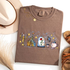 May include: A brown t-shirt with a graphic print of a castle, a rose in a glass dome, a teapot, a cup, and a candlestick. The graphic is surrounded by stars and a blue and white splatter pattern.