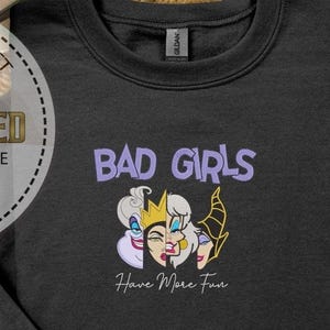 Puede incluir: Sudadera negra con el texto "BAD GIRLS" en morado sobre un gr&aacute;fico de tres caras de villanas de dibujos animados. Debajo, la frase "Have More Fun" en cursiva. La sudadera es de la marca Gildan.