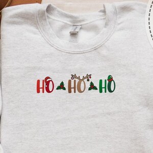 Może przedstawiać: Jasnoszara bluza z wyszytym napisem "HO HO HO" w kolorach czerwonym, brązowym i zielonym. Litery ozdobione są motywami świątecznymi, w tym czapką Świętego Mikołaja, porożem renifera i czapką elfa.