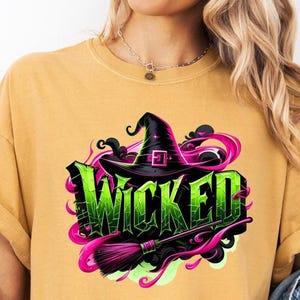 May include: Mustard yellow t-shirt featuring a graphic design with the word "Wicked" in green, a black witch hat, and a pink and black broom. The design includes pink and black swirls. The shirt is casual wear.
