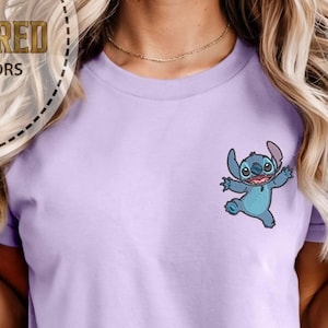 May include: A light purple t-shirt featuring a small embroidered image of the character Stitch from the animated film Lilo & Stitch. The cartoon character is blue with large eyes and is in a playful pose. The shirt has short sleeves.