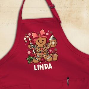 May include: A red apron with a gingerbread person graphic holding a candy cane and coffee cup. The gingerbread person is decorated with lights and a pink bow. The name "LINDA" is printed in white below the graphic. The apron has two pockets.