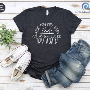 May include: A dark heather grey t-shirt with a white graphic of a sunrise with a heart in the center and the text "The sun will rise & And we will try again".
