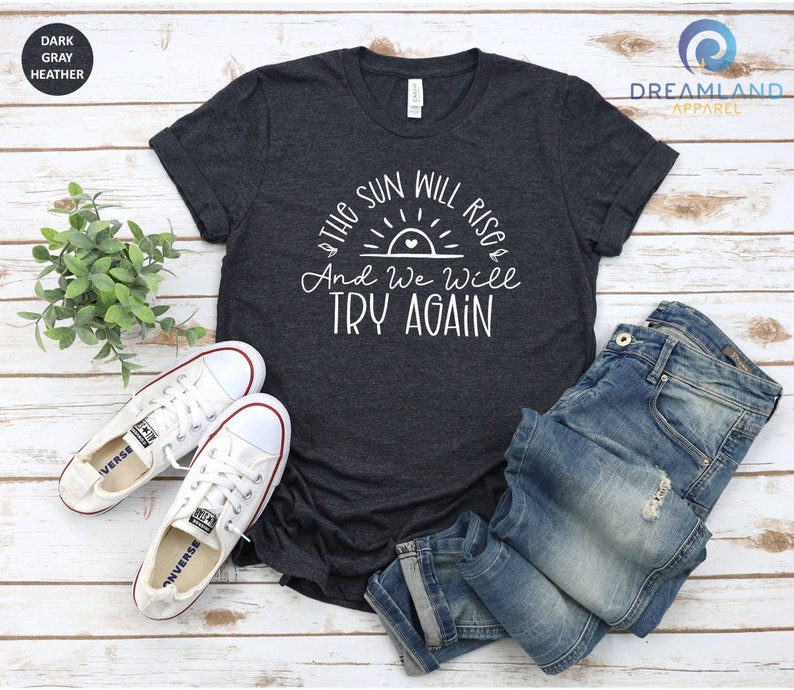 May include: A dark heather grey t-shirt with a white graphic of a sunrise with a heart in the center and the text "The sun will rise & And we will try again".