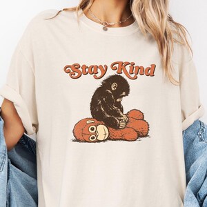 May include: Cream-colored t-shirt with the text "Stay Kind" in orange script. The graphic features a brown monkey sitting on a red teddy bear. The shirt has a relaxed fit.