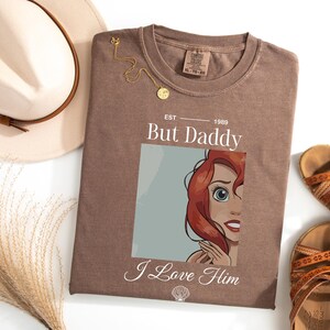 Może przedstawiać: Brązowa koszulka z napisem "But Daddy I Love Him" i ilustracją rudowłosej postaci z kresk&oacute;wki. Na zdjęciu znajduje się r&oacute;wnież złoty naszyjnik i beżowy kapelusz z brązową opaską. Obok koszulki znajdują się brązowe sandały.