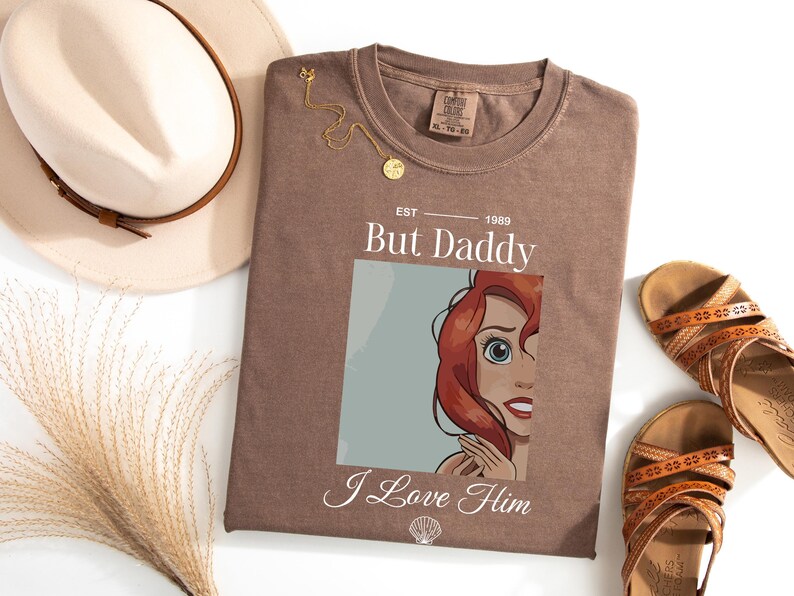 Może przedstawiać: Brązowa koszulka z napisem "But Daddy I Love Him" i ilustracją rudowłosej postaci z kresk&oacute;wki. Na zdjęciu znajduje się r&oacute;wnież złoty naszyjnik i beżowy kapelusz z brązową opaską. Obok koszulki znajdują się brązowe sandały.