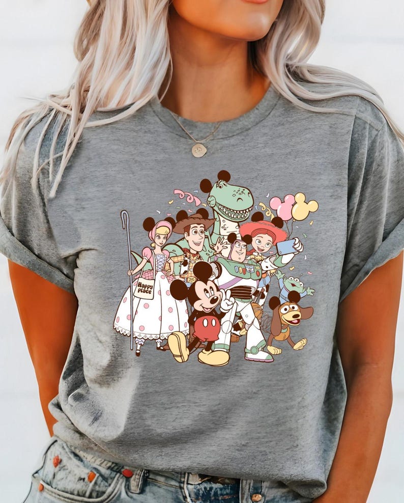 May include: Gray t-shirt featuring a colorful graphic of Disney characters, including Mickey Mouse, Buzz Lightyear, and Woody. The design includes balloons and confetti, creating a festive and playful vibe. The shirt is made of a soft material.