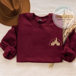 May include: A maroon sweatshirt with a gold castle graphic. The sweatshirt has long sleeves and a crew neck. A brown hat and decorative wheat stalks are in the background. The text "PERSONALIZED GIFT EMBROIDERED SWEATSHIRT & HOODIE" is in a circle.