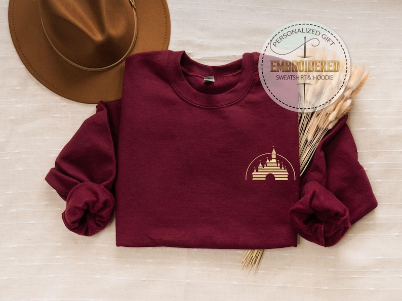 May include: A maroon sweatshirt with a gold castle graphic. The sweatshirt has long sleeves and a crew neck. A brown hat and decorative wheat stalks are in the background. The text "PERSONALIZED GIFT EMBROIDERED SWEATSHIRT & HOODIE" is in a circle.